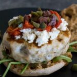 Open faced Mediterranean turkey burger topped with crumbled feta, chopped olives, and red peppers, served on a dark plate with arugula.