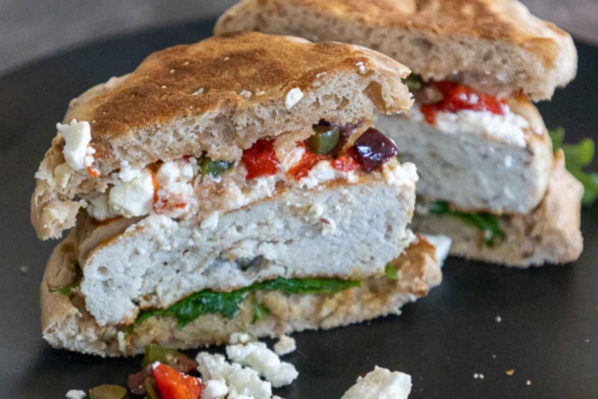 Sliced burger on a whole grain bun topped with fetat cheese, olives, roasted red peppers, and greens.