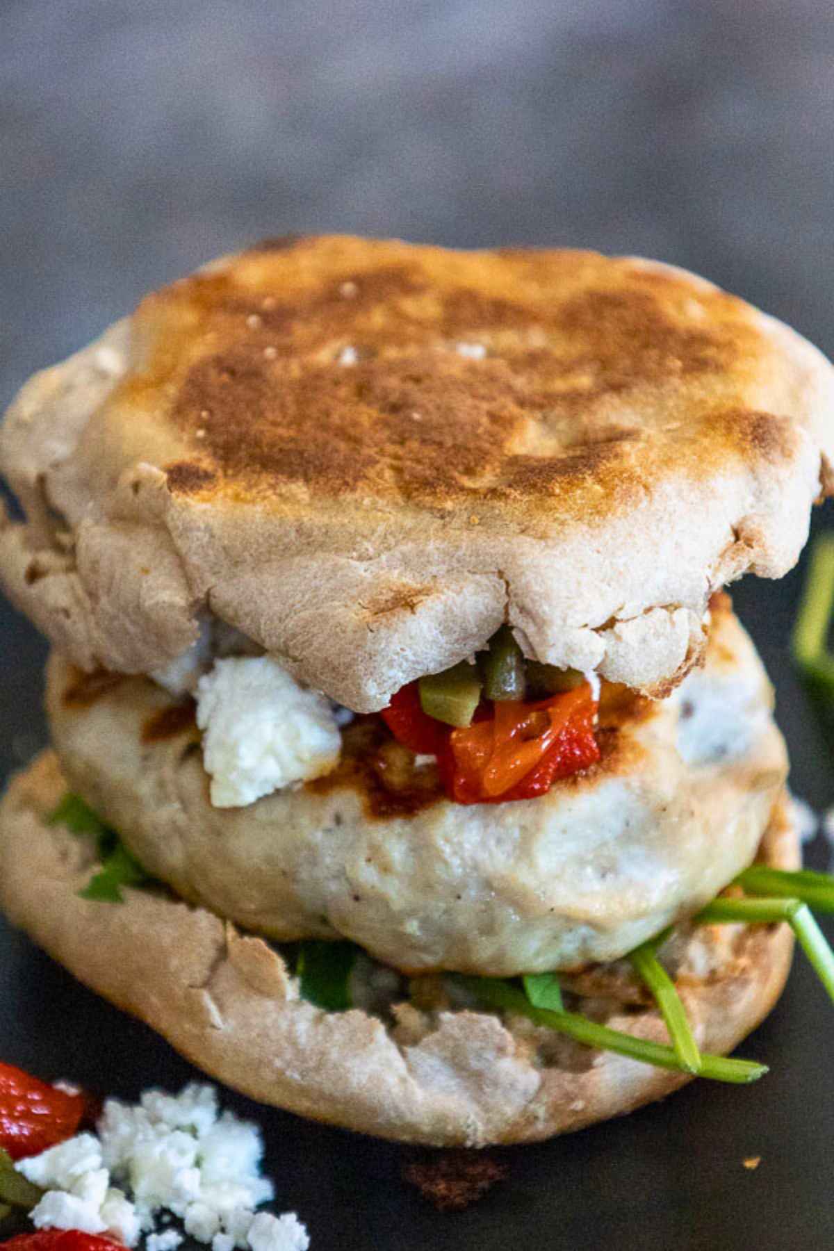 Whole Mediterranean turkey burger stacked with arugula, grilled turkey patty, feta cheese, olives, and red peppers on a toasted english muffin.