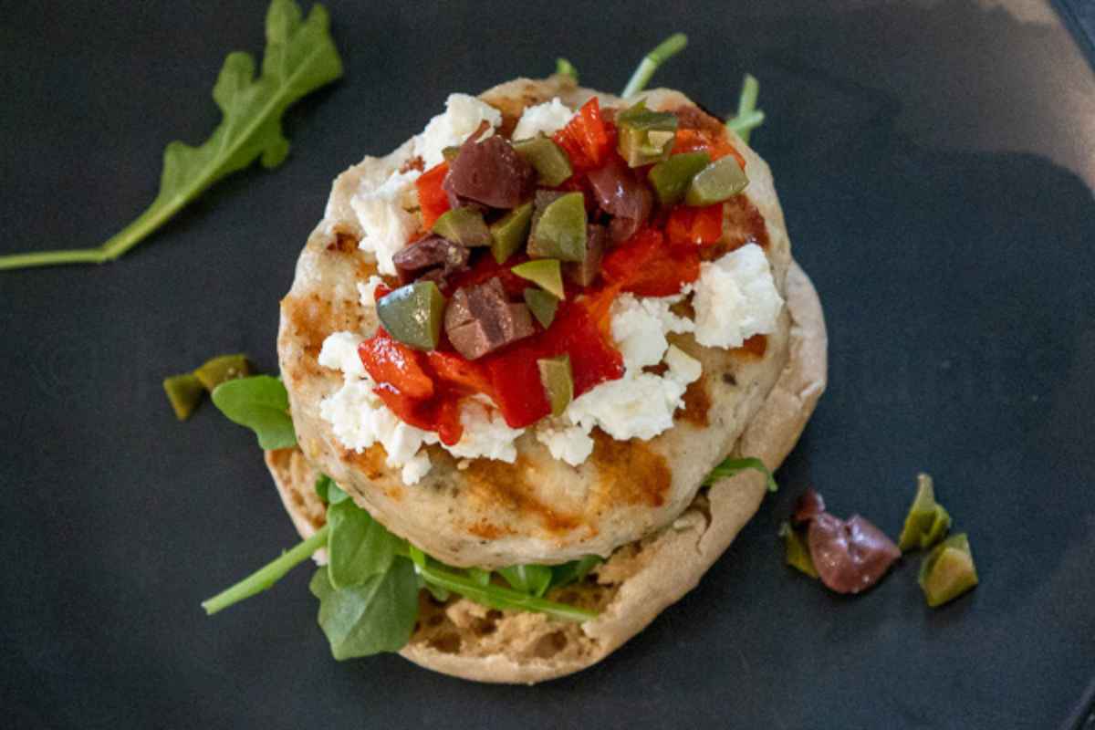 Overhead image of Open faced burger topped with crumbled feta, chopped olives, and red peppers, served on a dark plate with arugula.