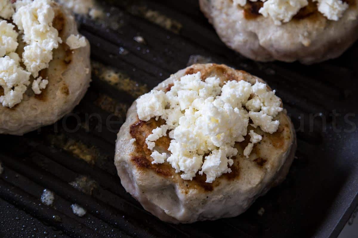Grilled turkey burgers topped with crumbled feta cheese cooking on a grill pan, with visible browning on the patty surface.