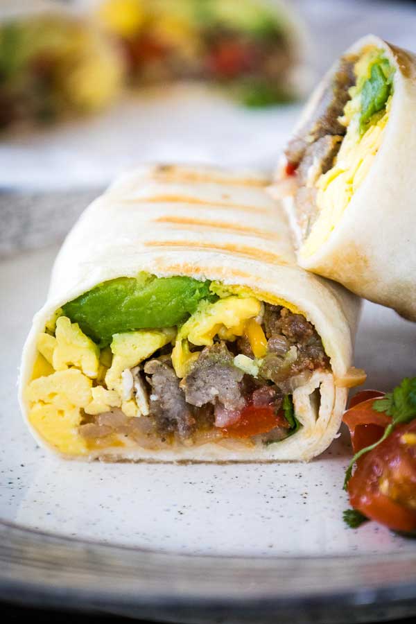 A close up of a breakfast burrito cut in half, showing scrambled eggs, avocado, sausage, cheese, and tomato inside a grilled tortilla.