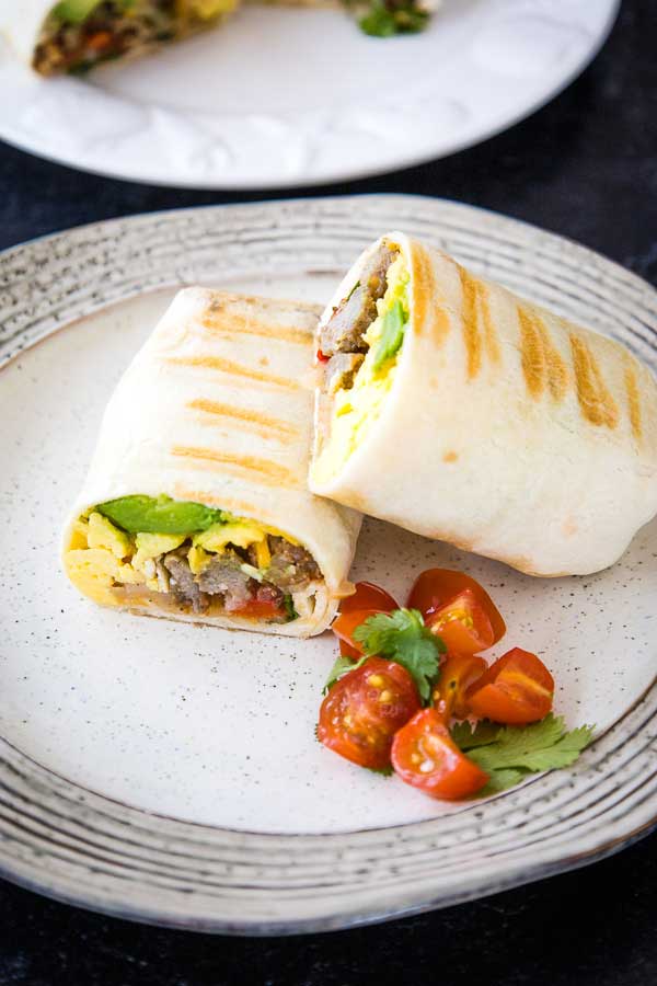 Two halves of a grilled breakfast burrito on a plate, served with cherry tomato halves and a sprig of fresh cilantro.