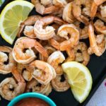 Close up of a platter of cooked peeled shrimp garnished with lemon wedges, served on a dark tray with a colorful napkin underneath.