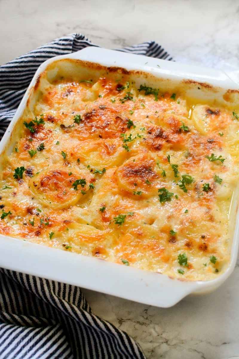 Overhead view of a fully baked scalloped potato casserole with a golden, bubbly cheese and parsley in a white baking dish on a striped towel.