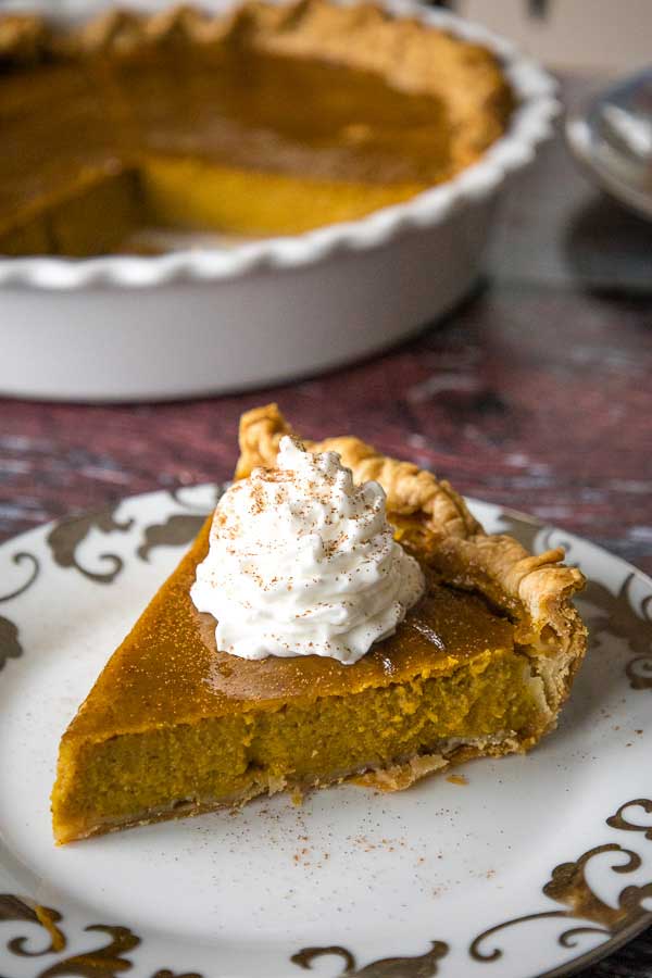 A slice of Maple Pumpkin Pie on a decorative plate topped with a swirl of whipped cream, with the rest of the pie in the background.