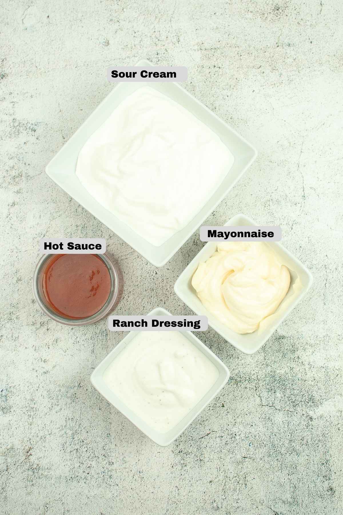 Overhead view of labeled ingredients in small bowls; sour cream mayonnaise, ranch dressing, and hot sauce on a light textured surface.