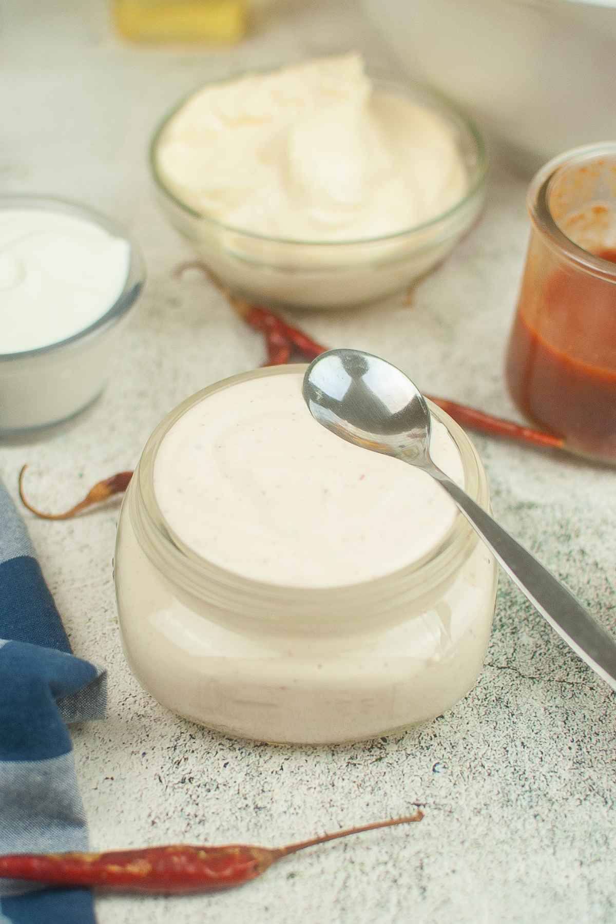 A small glass jar filled with spicy ranchero sauce with a spoon resting on top, surrounded by other ingredient bowls.