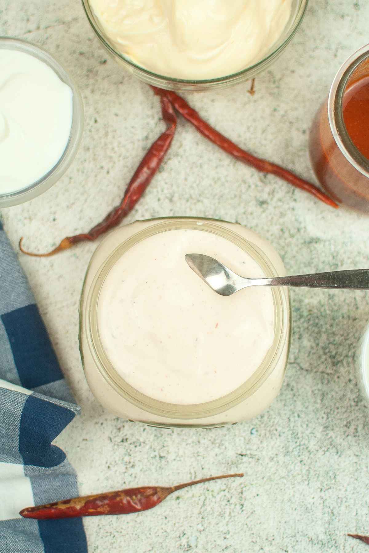 Overhead view of a glass jar filled with creamy ranchero sauce with a spoon resting on top, surrounded by bowls of ingredients and dried red chili peppers on a light textured surface.