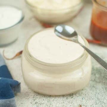 A small glass jar filled with spicy ranchero sauce with a spoon resting on top, surrounded by other ingredient bowls.