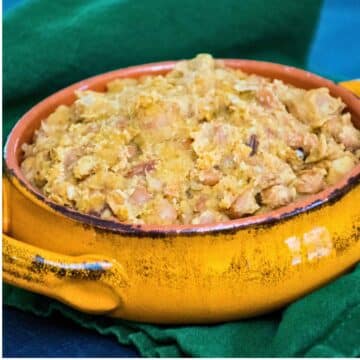 Close up of a rustic yellow ceramic bowl filled with homemade refried beans, set on a green cloth with a blue background.
