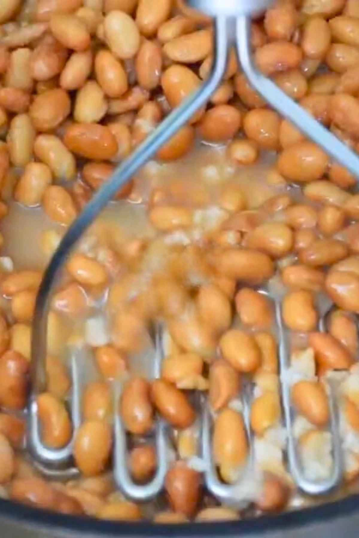 Cooked pinto beans being mashed wtih a potato masher in a pot.