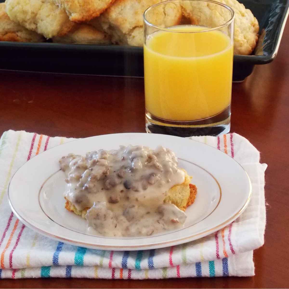 A biscuit topped with creamy sausage gravy on a white plate, set on a striped cloth napkin, with a glass of orange juice and a tray of biscuits in the background.