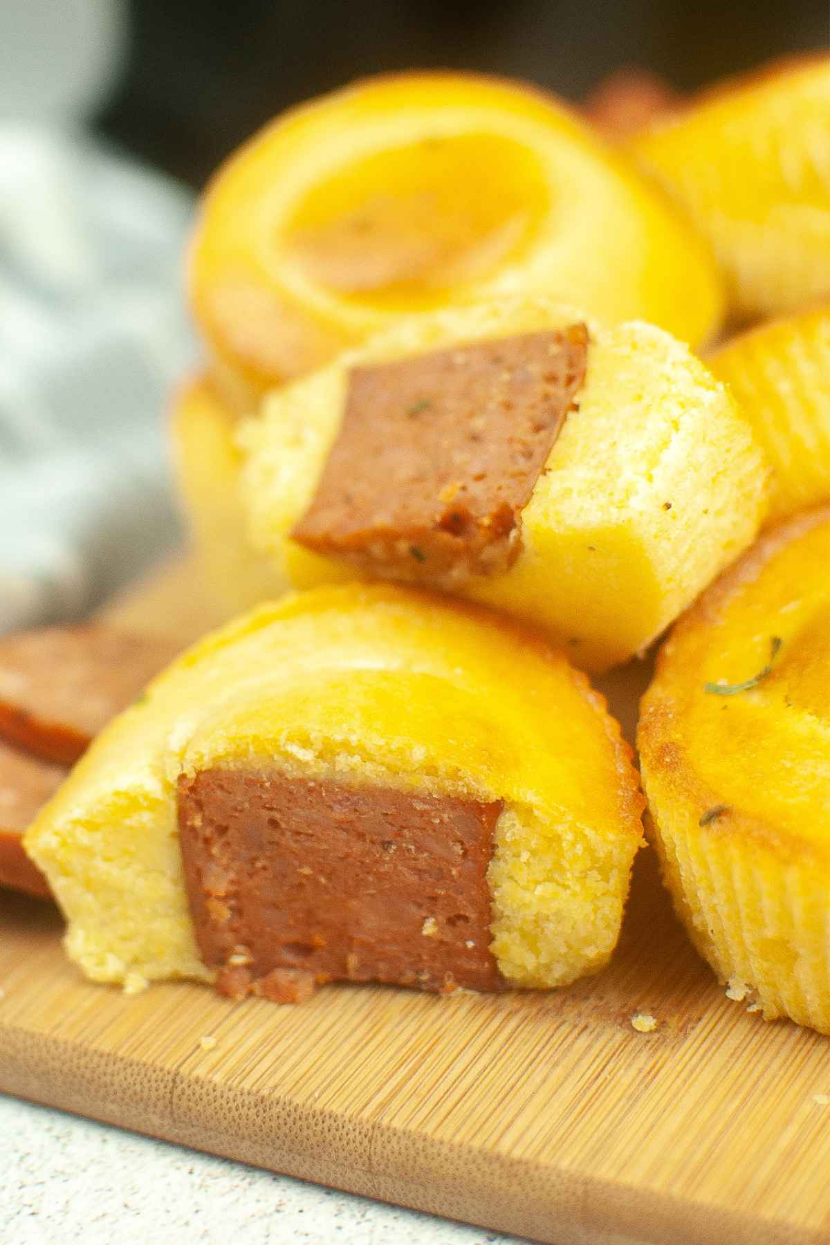 Upclose image with golden brown cornbread muffins, some with visible sausage centers resembling mini sausage corn dog bites, with sliced sausage scattered around.