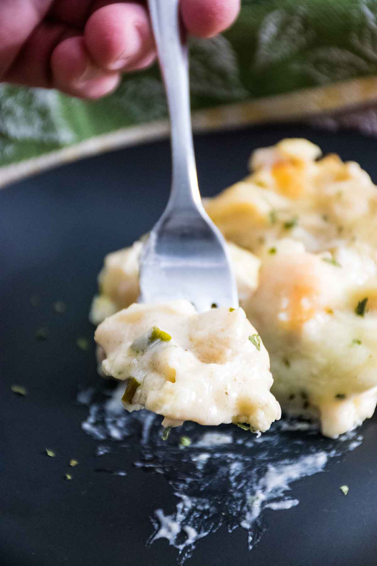 A close up of a fork lifting a creamy bite of seafood lasagna from a black plate, showing pieces of seafood and herbs in a rich white sauce.