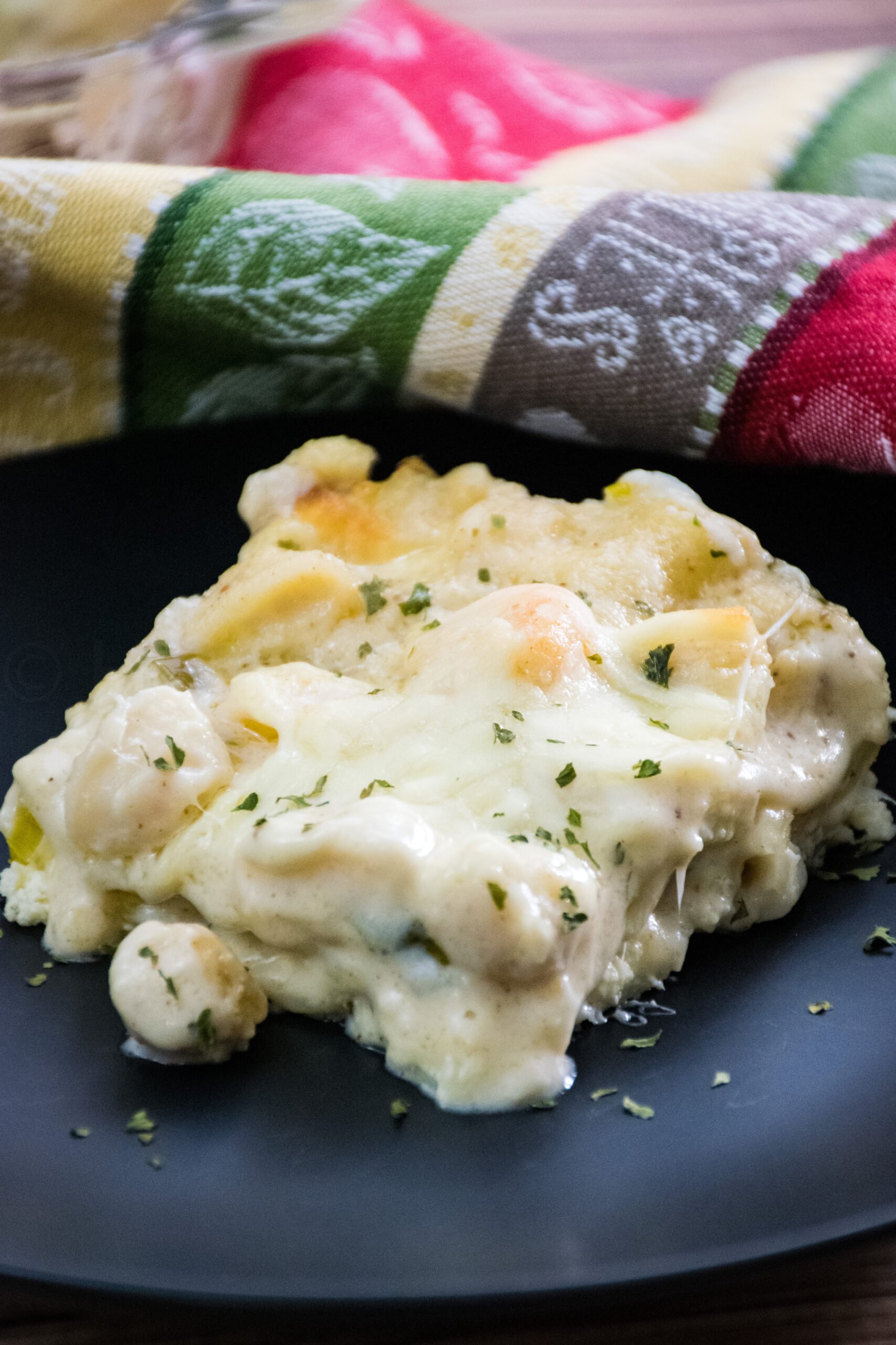 A slice of baked seafood lasagna served on a black plate, topped with melted cheese and garnished with dried herbs, with a colorful napkin in the background.
