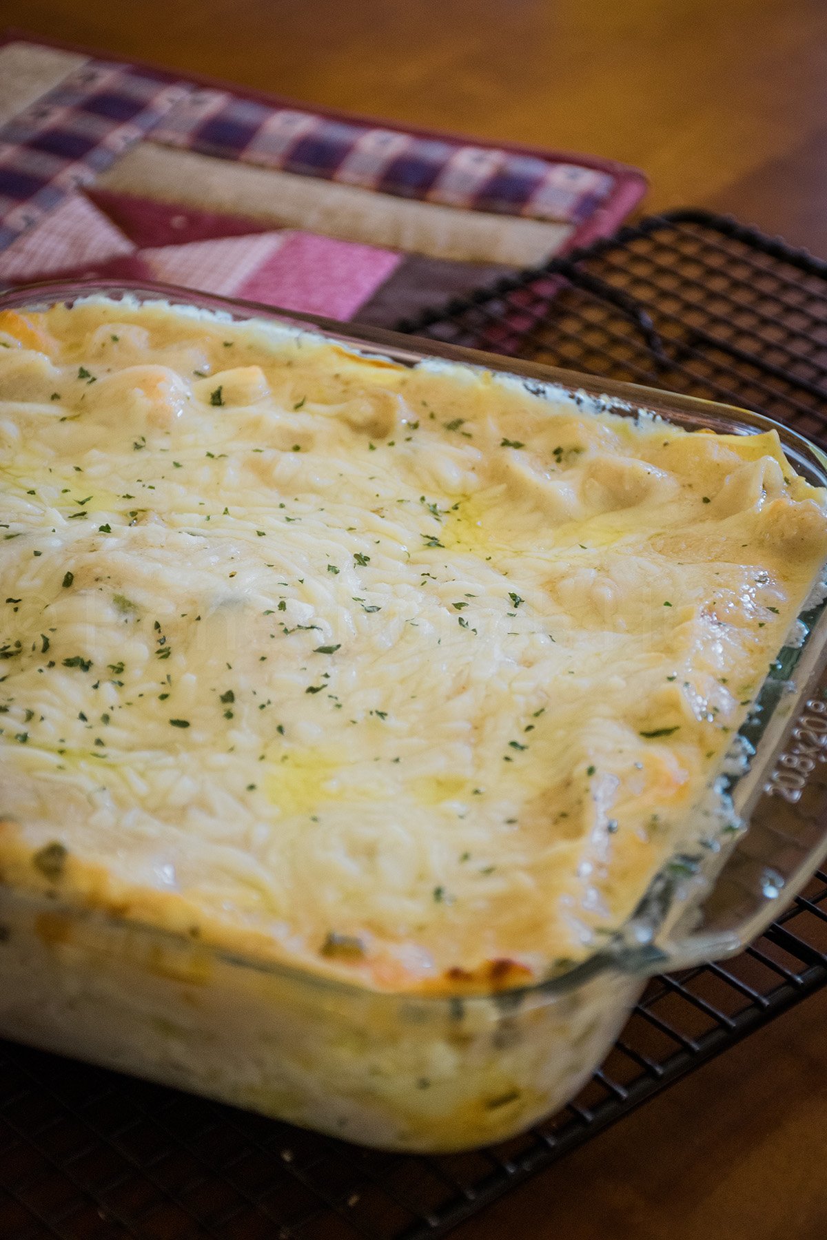 A freshly baked seafood lasagna in a glass baking dish, topped with melted cheese and sprinkled with herbs, resting on a cooling rack beside a quilted pot holder.
