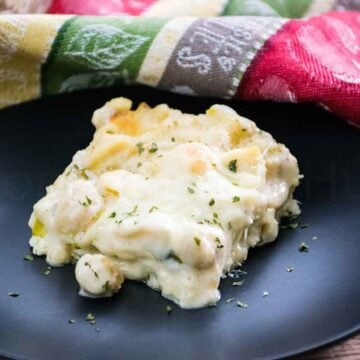 A slice of baked seafood lasagna served on a black plate, topped with melted cheese and garnished with dried herbs, with a colorful napkin in the background.
