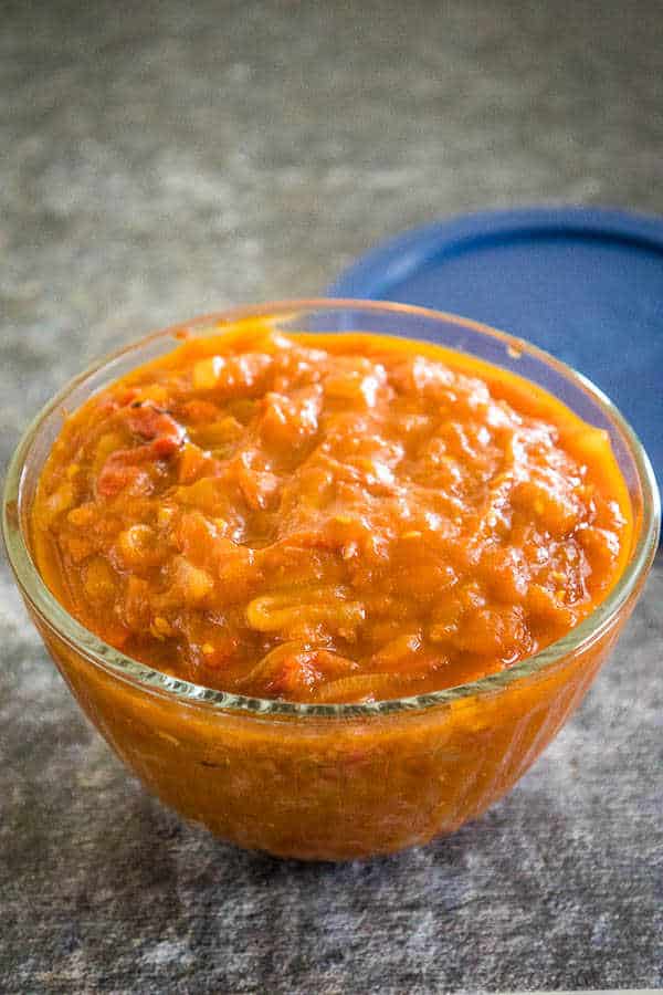 Side angle of a glass bowl filled with vibrant orange-red spicy tomato chutney, set on a gray surface with a blue lid in the background.