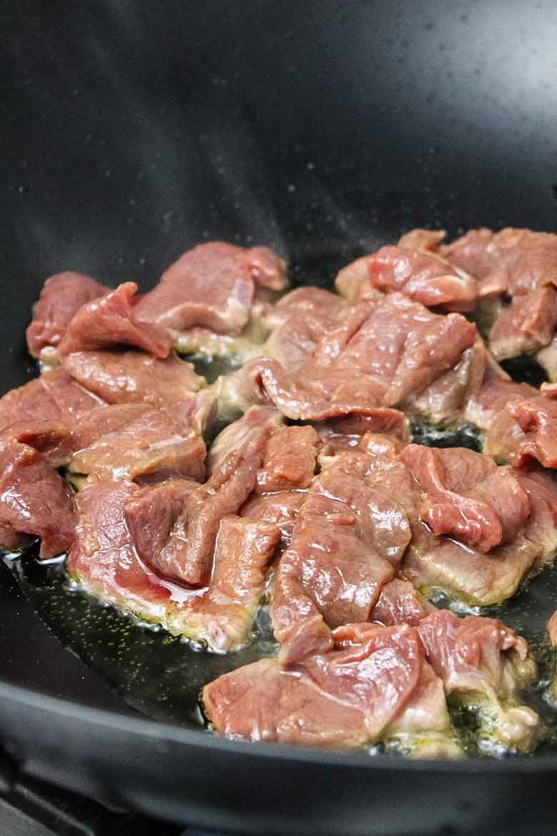 Raw slices of venison cooking in a hot skillet with oil, starting to brown around the edges.