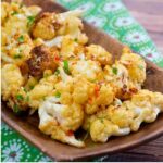 Close up of golden brown roasted cauliflower florets garnished with chopped cilantro, served on a wooden tray placed over a green floral-patterned cloth.