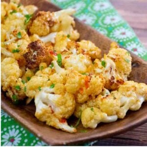 Close up of golden brown roasted cauliflower florets garnished with chopped cilantro, served on a wooden tray placed over a green floral-patterned cloth.