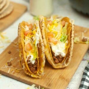 Two cheesy gordita crunch tacos filled with seasoned meat, lettuce, shredded cheese, diced tomatoes, and sour cream on a wooden board.