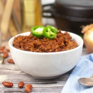 A white bowl filled with thick, hearty chili tiopped with fresh slices of jalapeno pepper, set on a rustic wooden surface with scattered beans and a blue napkin.