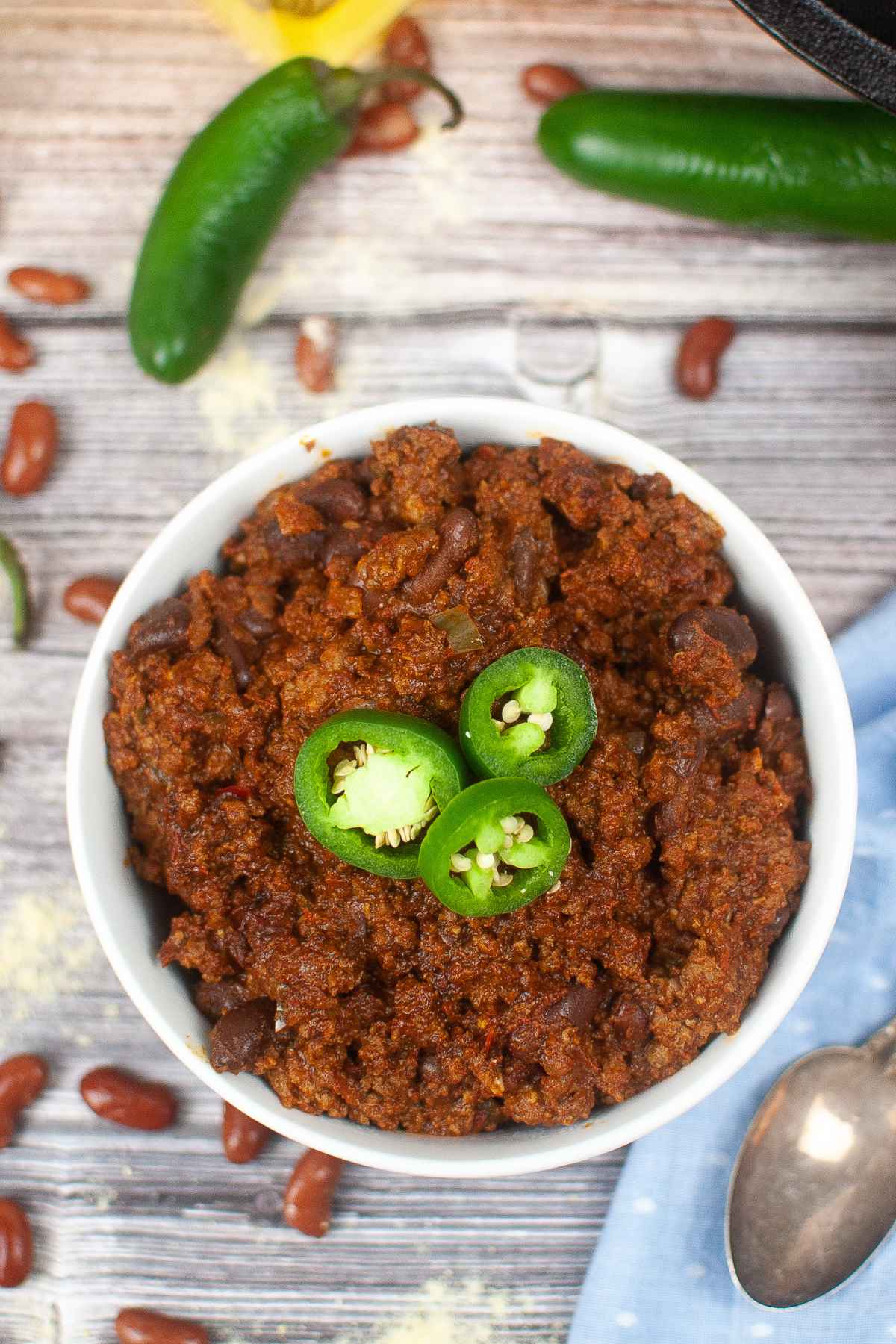 Overhead shot of a bowl of chili garnished with sliced jalapenos, surrounded by dried beans and whole jalapeno peppers on a rustic wooden table.