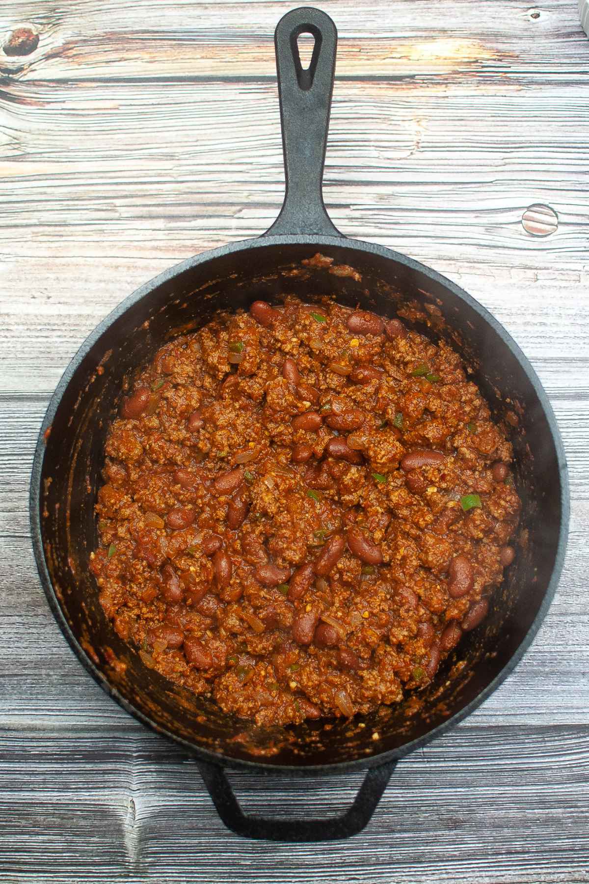 Overhead view of a cast iron skillet filled with chunky Texas Roadhouse Chili made with ground meat, beans, and spices on a wood grained background.