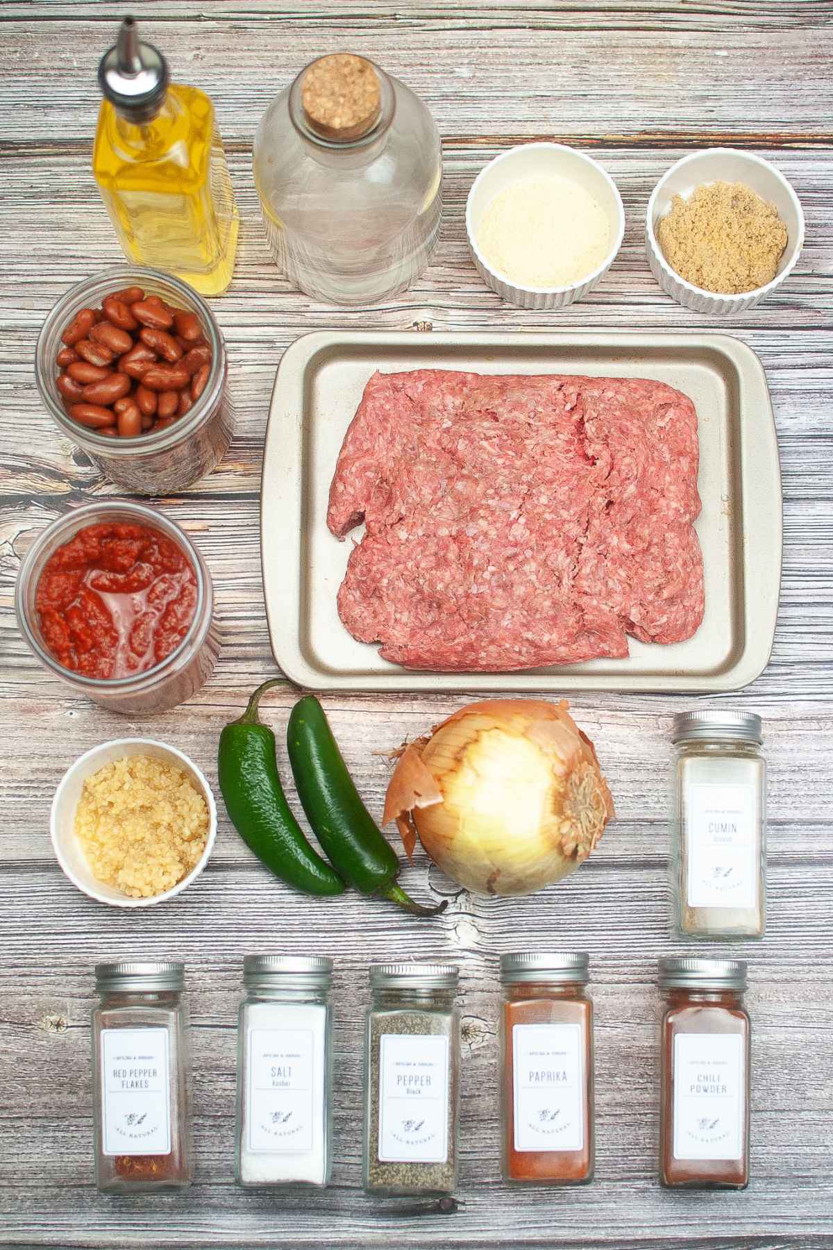 Flat lay of raw ingredients for chili including ground meat, canned beans, tomatoes, spices, onion, garlic, jalapenos, and bottles of oil and vinegar arranged on a wooden surface.