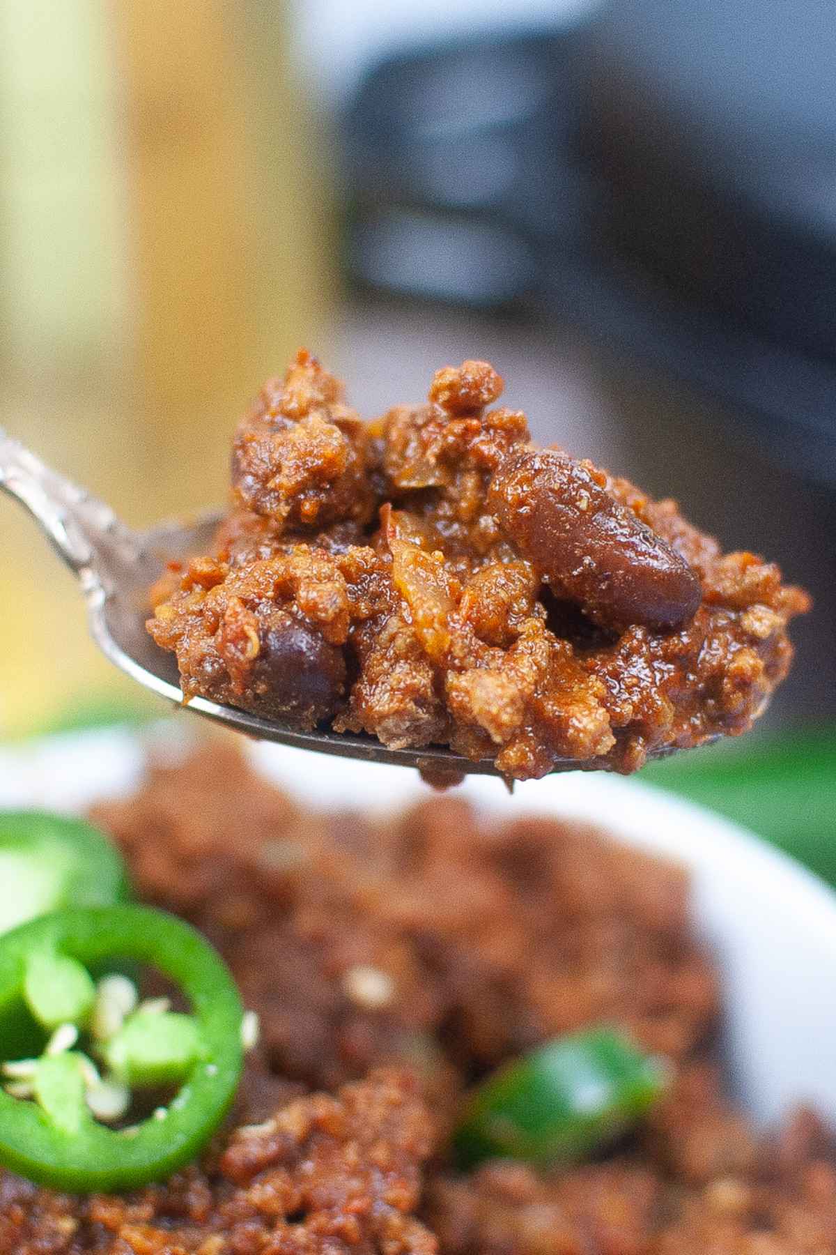 Close up of a spoonful of meaty chili with beans and visible spices above a bowl, showcasing the rich texture and vibrant color.