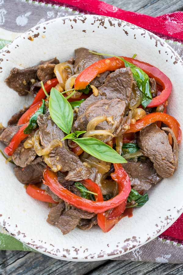 Overhead view of Thai basil Venison stir fry in a rustic white bowl, featuring slices of venison, red bell pepper, onions, an fresh Thai basil leaves.