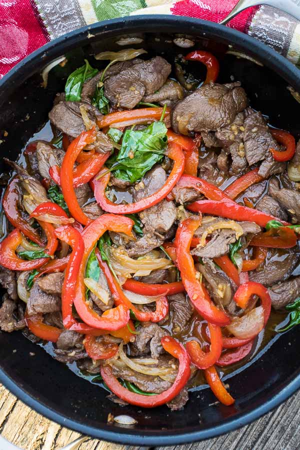 Close up of Thai basil venison stir fry in a skillet, showcasing sliced venison, red bell pepper strips, onion, and wilted Thai basil in a savory sauce.