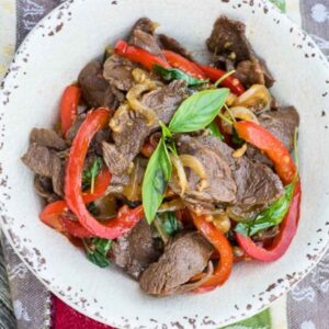 Overhead view of Thai basil Venison stir fry in a rustic white bowl, featuring slices of venison, red bell pepper, onions, an fresh Thai basil leaves.