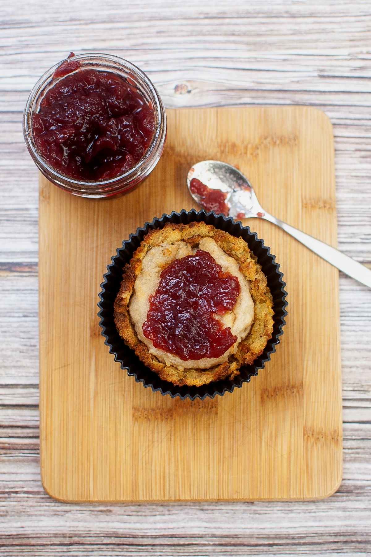 Overhead shot of stuffing muffin in silicone cup placed on a wooden board next to a small jar and spoon of cranberry sauce.