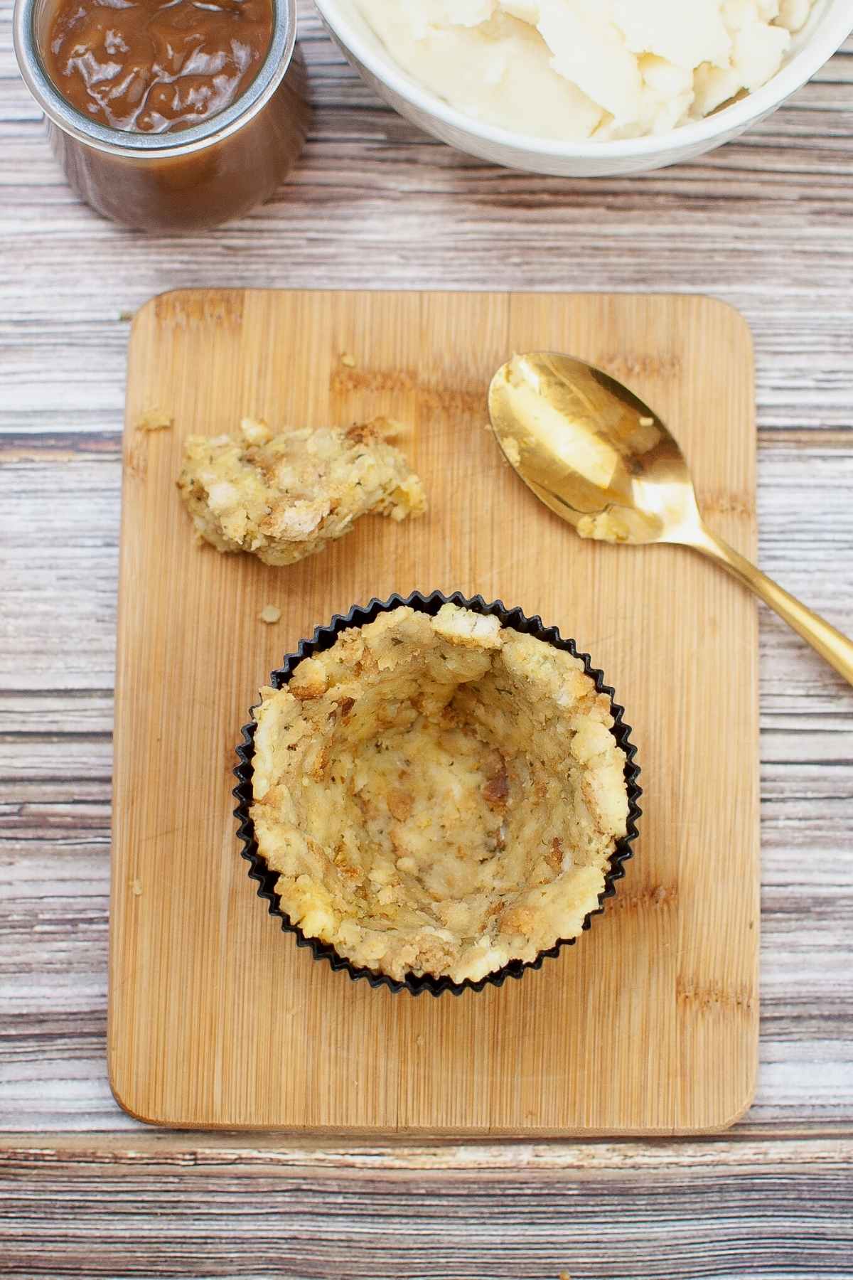 Unstuffed stuffing shell in silicone cup on a wooden board with mashed potatoes and gravy in bowls nearby, showing the hollowed center ready for filling.