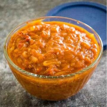 Side angle of a glass bowl filled with vibrant orange-red spicy tomato chutney, set on a gray surface with a blue lid in the background.