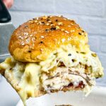A hot turkey cranberry slider being lifted on a spatula, with melted cheese, turkey, stuffing, and cranberry sauce inside a golden, seeded bun.