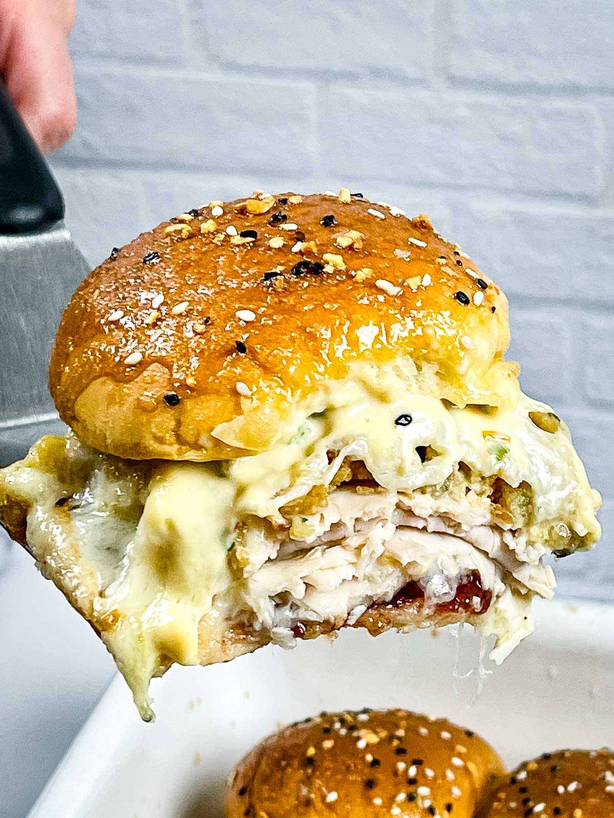 A hot turkey cranberry slider being lifted on a spatula, with melted cheese, turkey, stuffing, and cranberry sauce inside a golden, seeded bun.