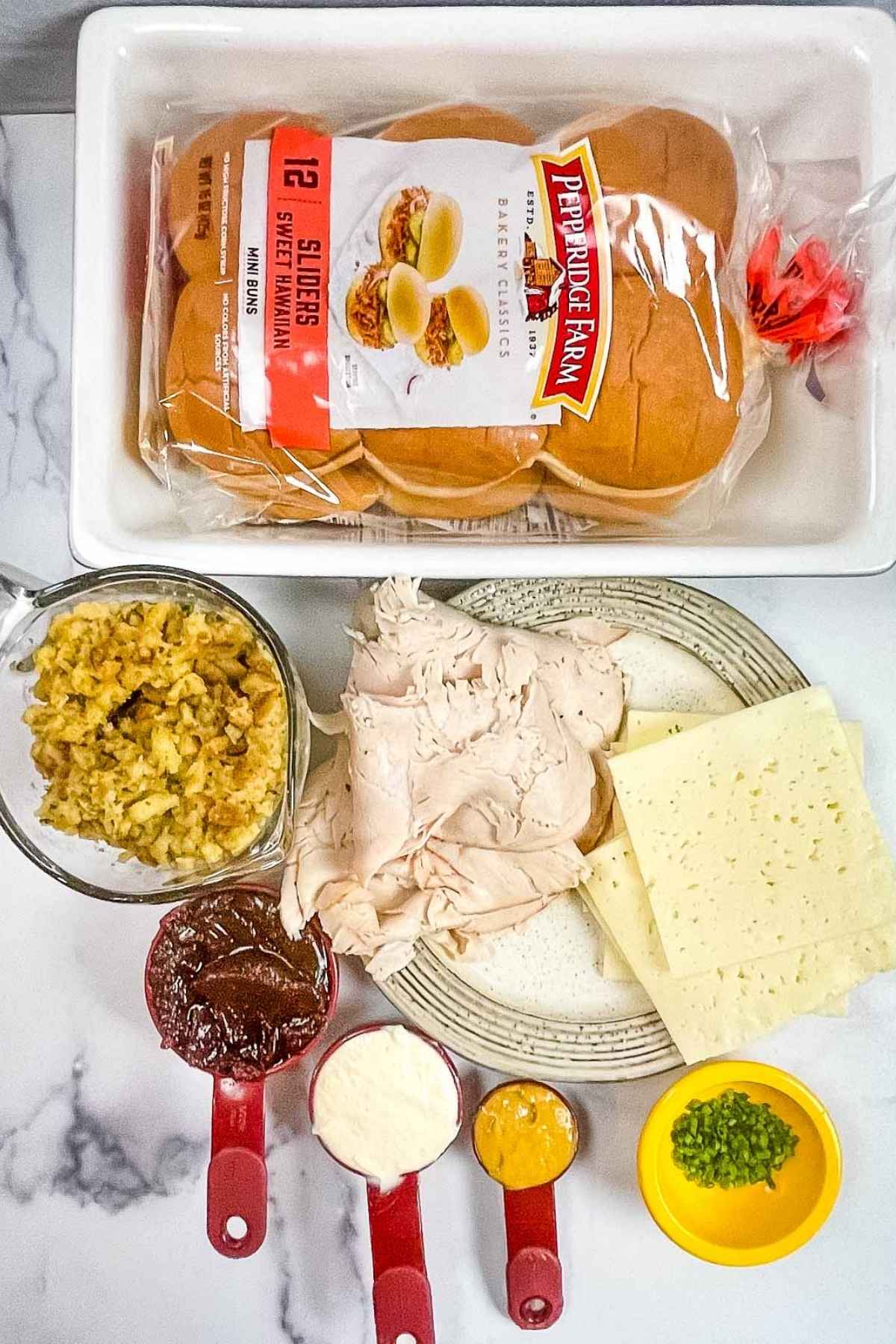 Overhead view of ingredients for slider recipe: Hawaiian rolls, turkey slices, swiss cheese, stuffing, cranberry sauce, mayo, mustard, and chopped green onions.