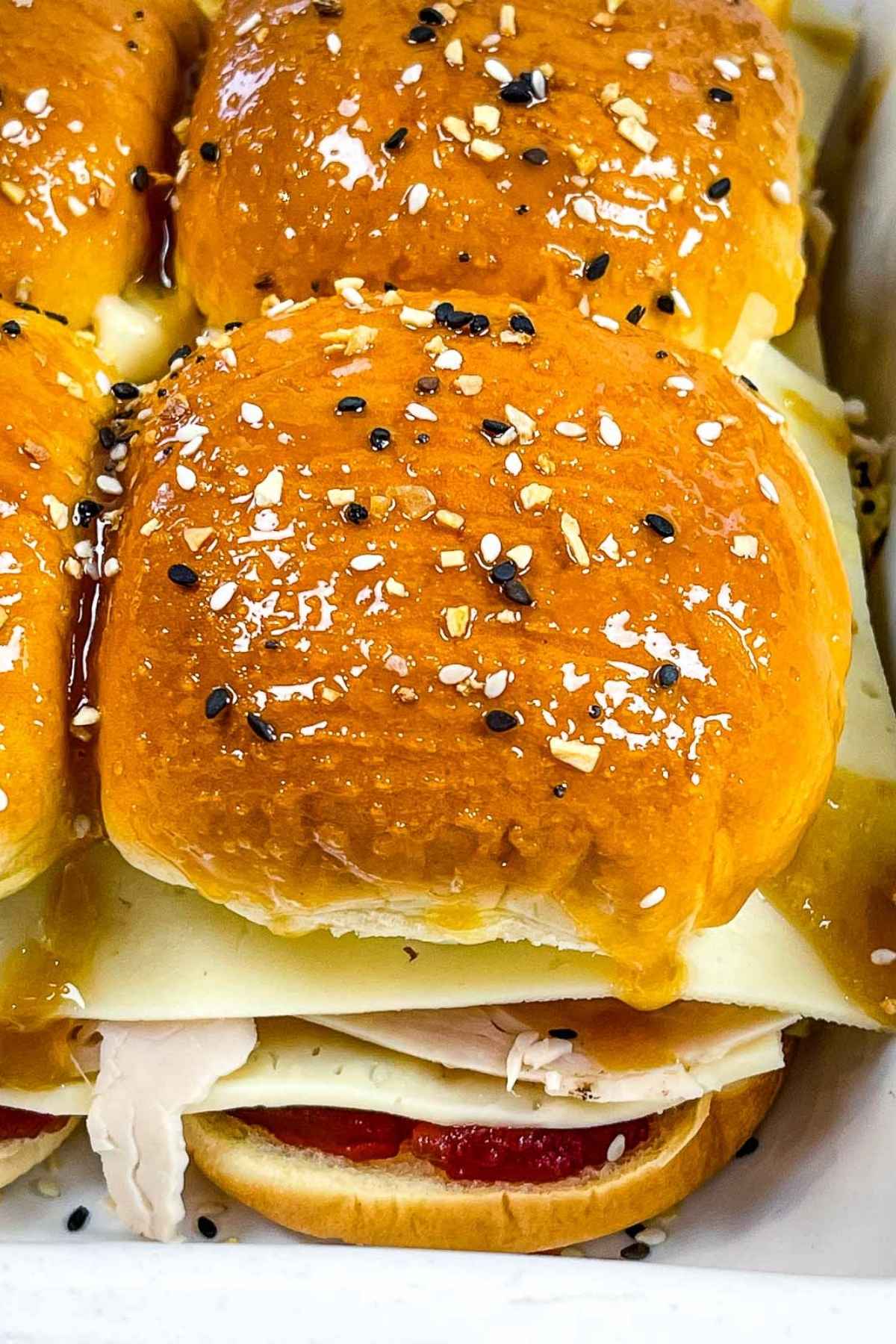 Close up of a turkey slider in a baking dish, showing layers of sliced turkey, cranberry sauce, and cheese on a sweet Hawaiian roll.
