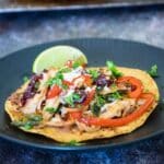 A turkey tostada topped with shredded turkey, sauteed red peppers, black beans, cheese, and chopped cilantro, served with a lime wedge on a dark plate