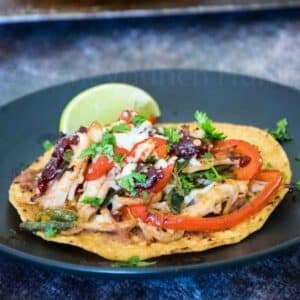 A turkey tostada topped with shredded turkey, sauteed red peppers, black beans, cheese, and chopped cilantro, served with a lime wedge on a dark plate