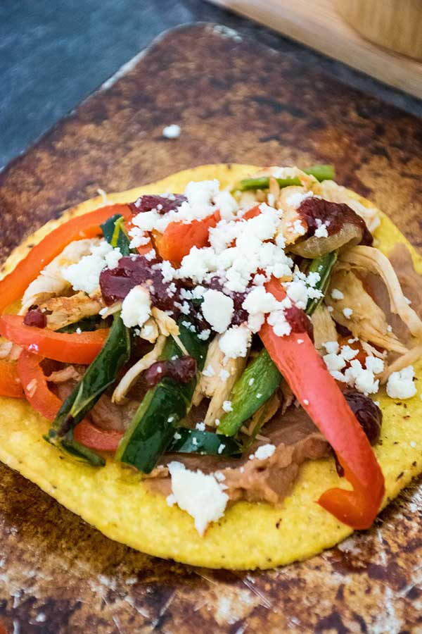 A close up of a turkey tostada layered with refried beans sauteed peppers, shredded turkey, onions, and crumbled cheese on a rustic baking sheet.