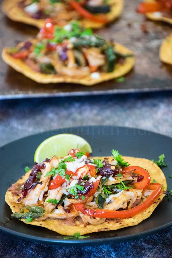 A turkey tostada topped with shredded turkey, sauteed red peppers, black beans, cheese, and chopped cilantro, served with a lime wedge on a dark plate wih a sheet pan of tostadas in the background.