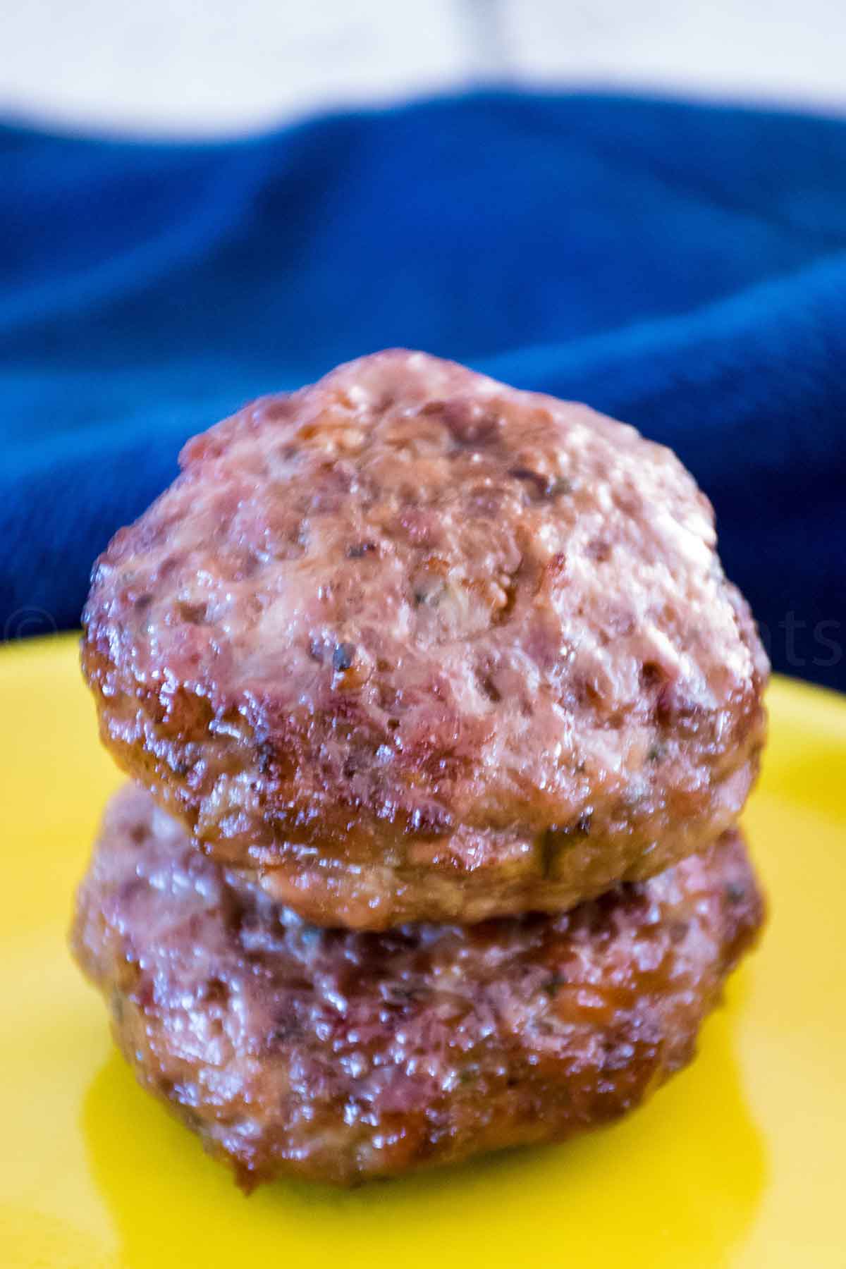 Two cooked venison breakfast sausage patties stacked on a yellow plate, with a blue cloth in the background.