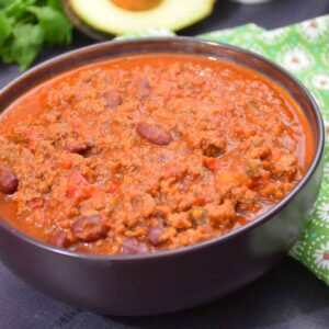 A close up of a bowl filled with chili made with ground venison and beans, with an chopped avocado in a white bowl in the back ground and an avocado half next to it.