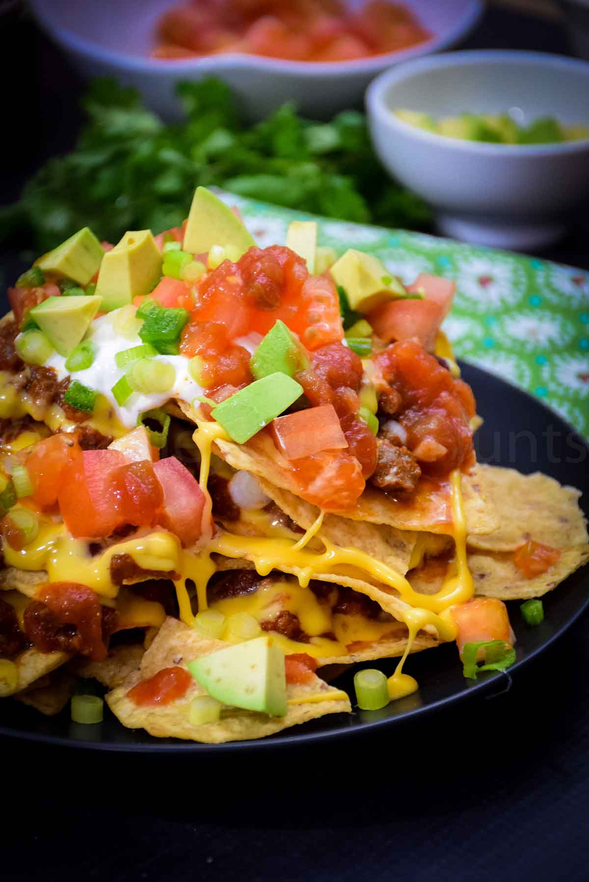 A heaping plate of loaded nachos piled high with melted cheese, ground meat, diced tomatoes, avocado chunks, salsa, sour cream, and sliced green onions, served on a black plate.