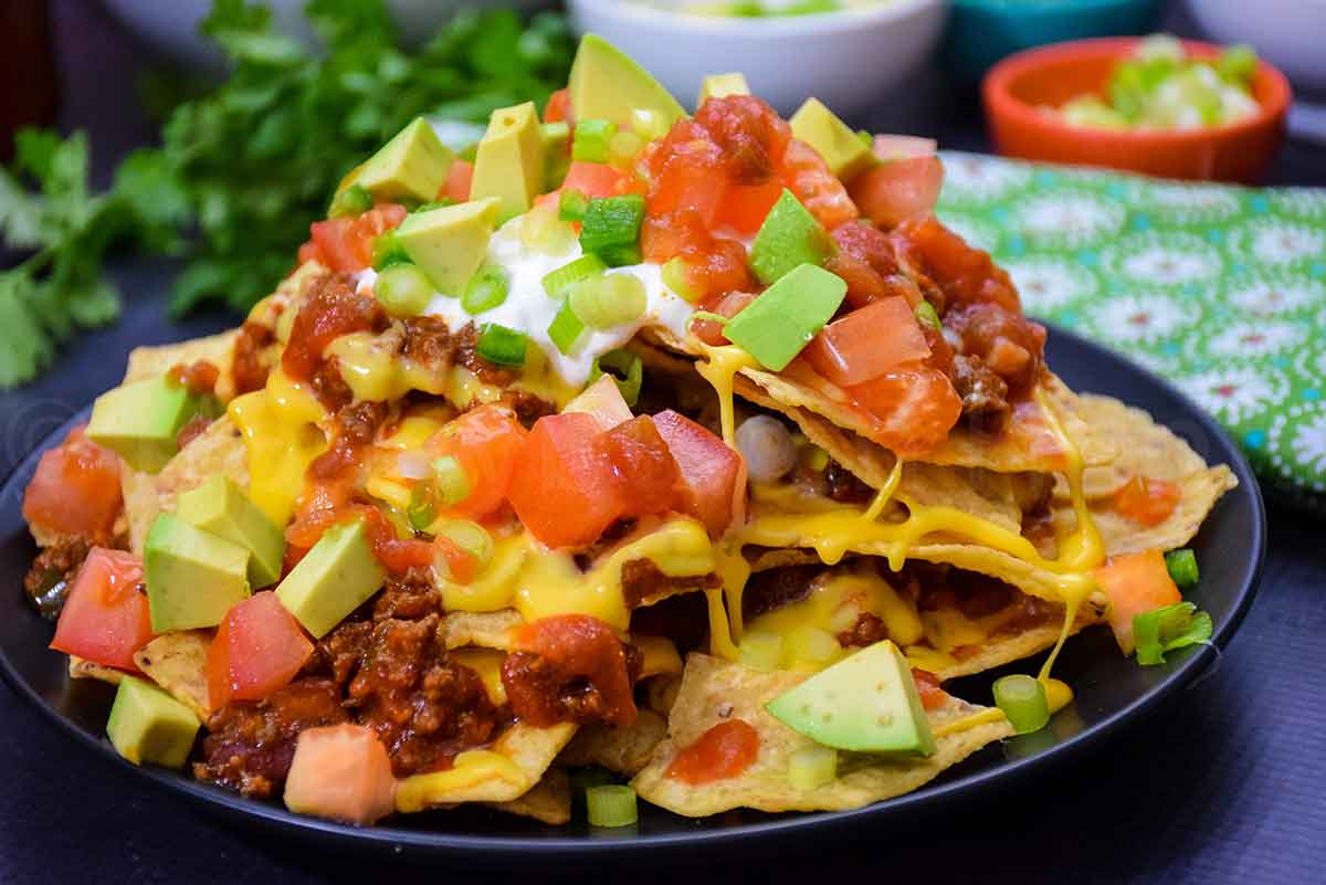 A heaping plate of loaded nachos piled high with melted cheese, ground meat, diced tomatoes, avocado chunks, salsa, sour cream, and sliced green onions, served on a black plate.